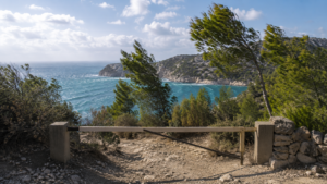Barrière fermée sur un sentier de garrigue menant à une calanque aux eaux turquoise, pins agités par le vent
