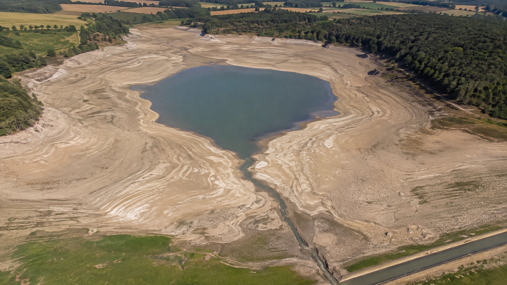 Vue aérienne d'un barrage-réservoir de Bourgogne à niveau très bas, large anneau de berges asséchées autour de l'eau et rigole partant vers le canal.