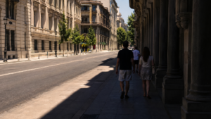 Rue de ville large à midi en été : l’ombre des immeubles couvre la chaussée et un trottoir, des passants vus de dos marchent dans la bande ombragée, le trottoir d’en face reste en plein soleil.