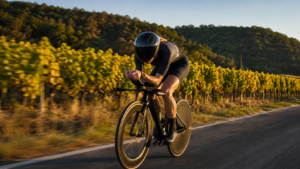 Cycliste anonyme en position aérodynamique sur un vélo de contre-la-montre, casque profilé à visière sombre, sur une route bordée de vignes.