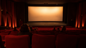 Salle de cinéma aux fauteuils rouges vue des derniers rangs, quelques spectateurs dispersés face à un écran allumé dans la pénombre.