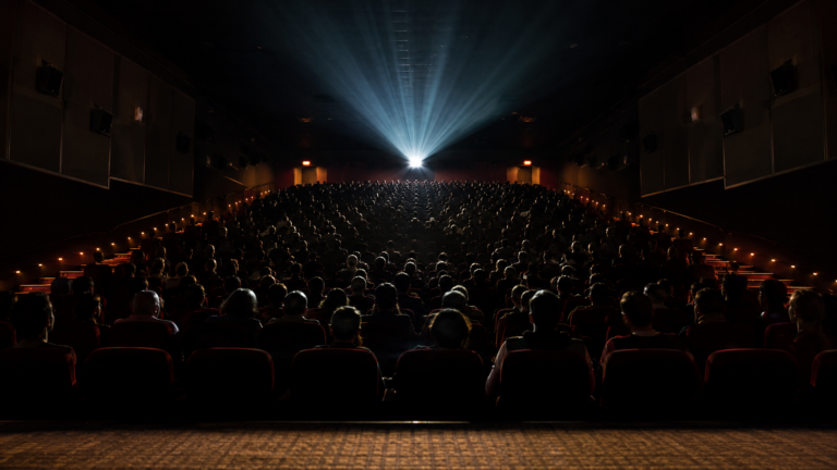 Salle de cinéma remplie vue depuis l'écran, faisceau du projecteur traversant l'obscurité, spectateurs en silhouette de dos.