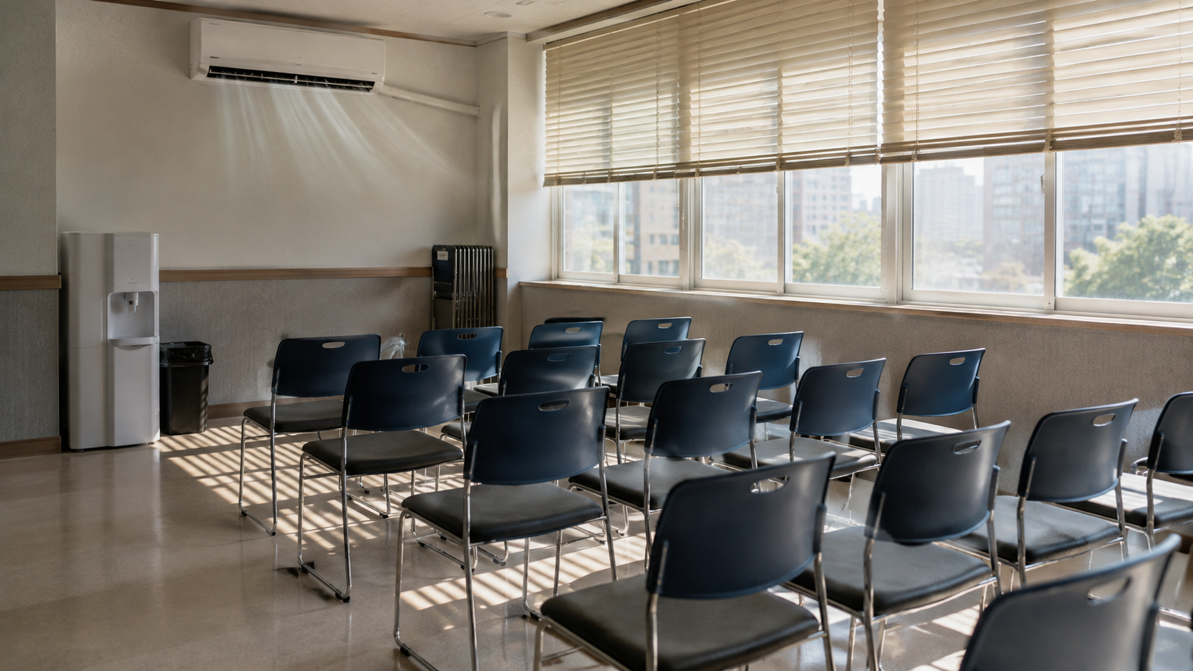 Salle de rafraîchissement public vide dans un centre communautaire : rangées de chaises, climatiseur mural, fontaine à eau, lumière filtrée par des stores à demi baissés.