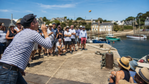 Sur un petit port de pêche breton, une foule joyeuse regarde un participant souffler un coquillage au-dessus d'une piste tracée sur le quai, par un après-midi d'été ensoleillé.