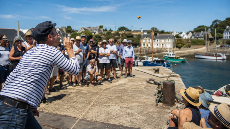 Sur un petit port de pêche breton, une foule joyeuse regarde un participant souffler un coquillage au-dessus d'une piste tracée sur le quai, par un après-midi d'été ensoleillé.