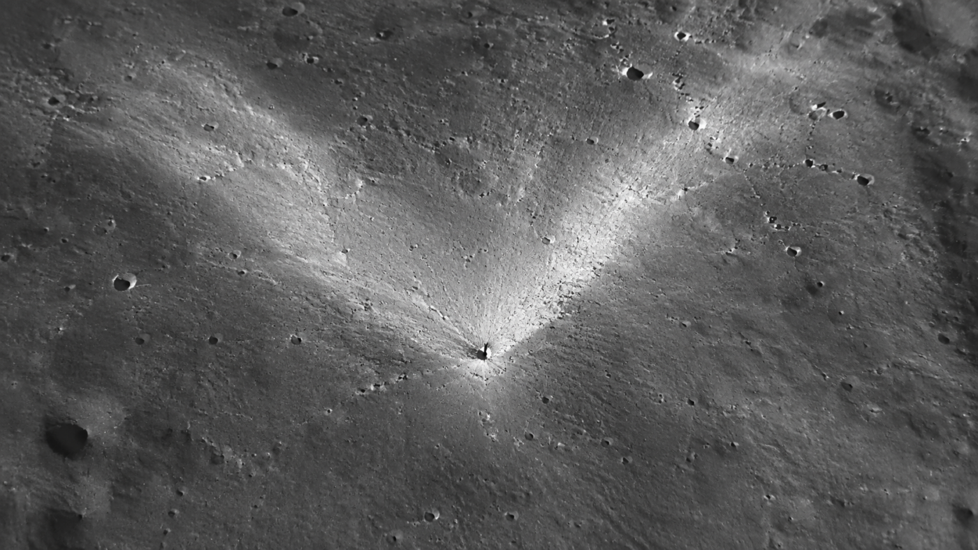Vue orbitale rapprochée de la surface lunaire grise, petit cratère d’impact récent entouré d’une gerbe d’éjectas clairs en forme de V, ombres allongées