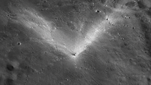 Vue orbitale rapprochée de la surface lunaire grise, petit cratère d’impact récent entouré d’une gerbe d’éjectas clairs en forme de V, ombres allongées