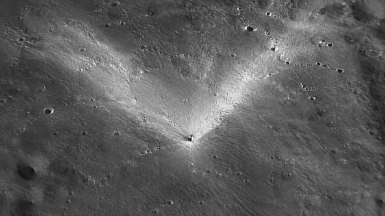 Vue orbitale rapprochée de la surface lunaire grise, petit cratère d’impact récent entouré d’une gerbe d’éjectas clairs en forme de V, ombres allongées