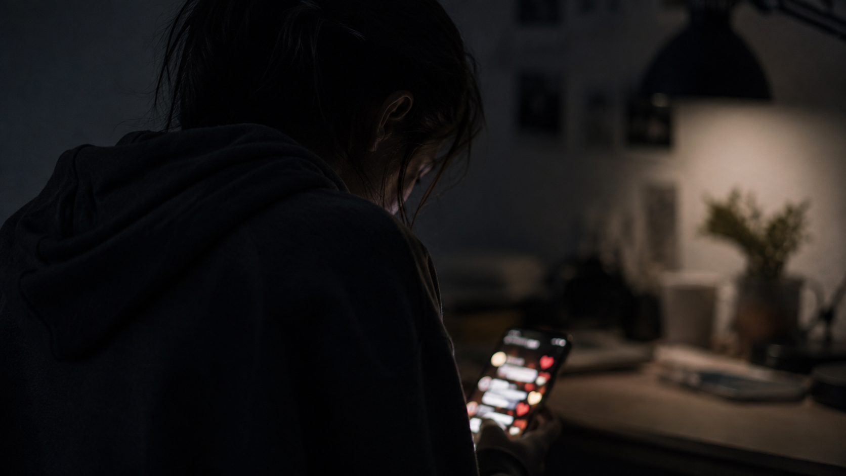 Des mains tenant un smartphone dans l’obscurité, l’écran lumineux affichant un flux flou de commentaires et d’icônes de cœur