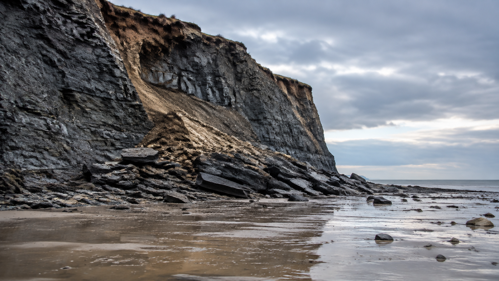 Falaise de marne sombre s’éboulant au-dessus d’un large estran à marée basse, blocs tombés et galets sur le sable mouillé