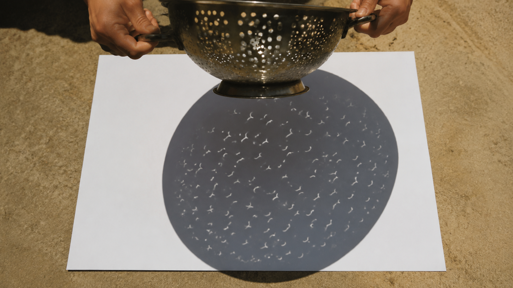 Deux mains tiennent une passoire de cuisine au-dessus d'une feuille blanche posée au sol, où se projettent des dizaines de petits croissants lumineux.