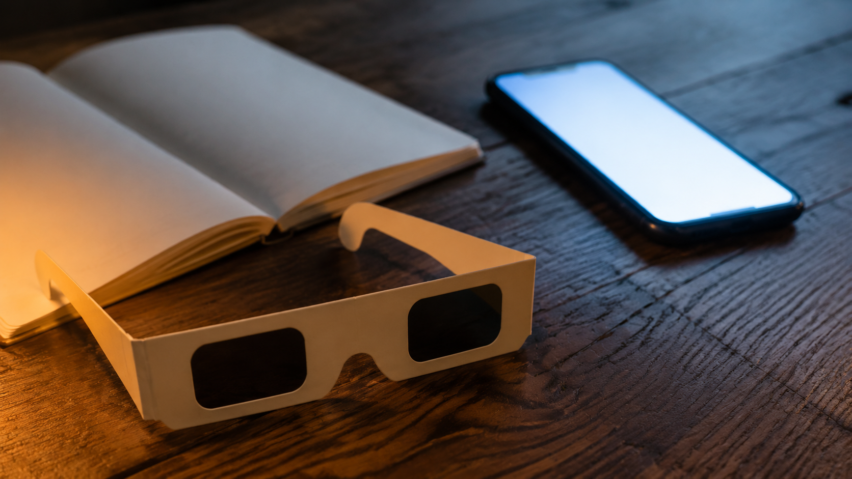 Lunettes d’éclipse en carton posées sur une table en bois à côté d’un téléphone à l’écran allumé et illisible et d’un carnet ouvert.