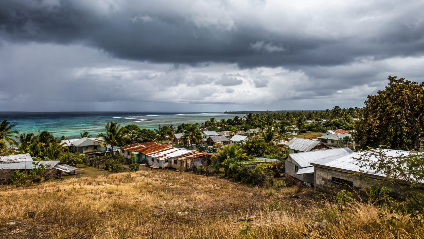 Village côtier du Pacifique sud aux toits de tôle, herbe sèche au premier plan sous un ciel chargé.