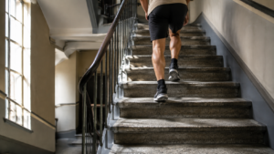 Une cage d’escalier d’immeuble éclairée par une haute fenêtre, des jambes en pleine montée aperçues de dos sur le palier.