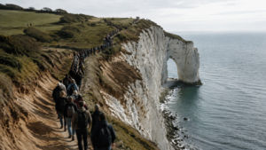 File de visiteurs sur un sentier étroit au sommet de falaises de craie blanches, herbe piétinée et sol érodé au bord du chemin, arche naturelle en arrière-plan.