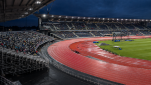 Stade d’athlétisme éclairé en soirée, piste rouge et tribunes démontables installées dans les deux virages, public vu de loin sans visage identifiable
