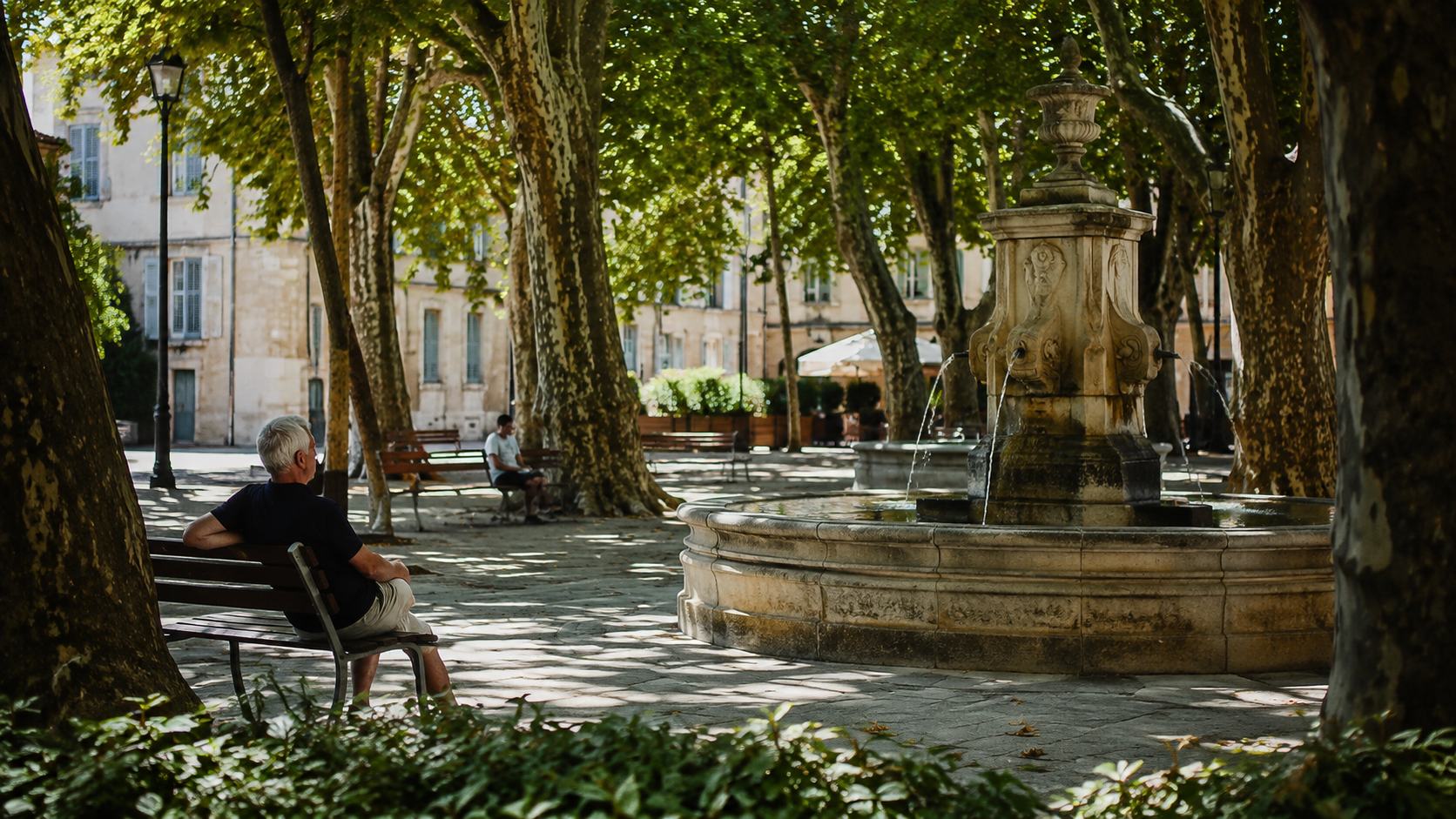Place ombragée par de grands platanes avec fontaine publique et bancs à l'ombre, dans une ville française.