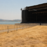 Site de festival désert au bord d'un lac, scène montée mais éteinte, aucun public, herbe sèche sous la chaleur.