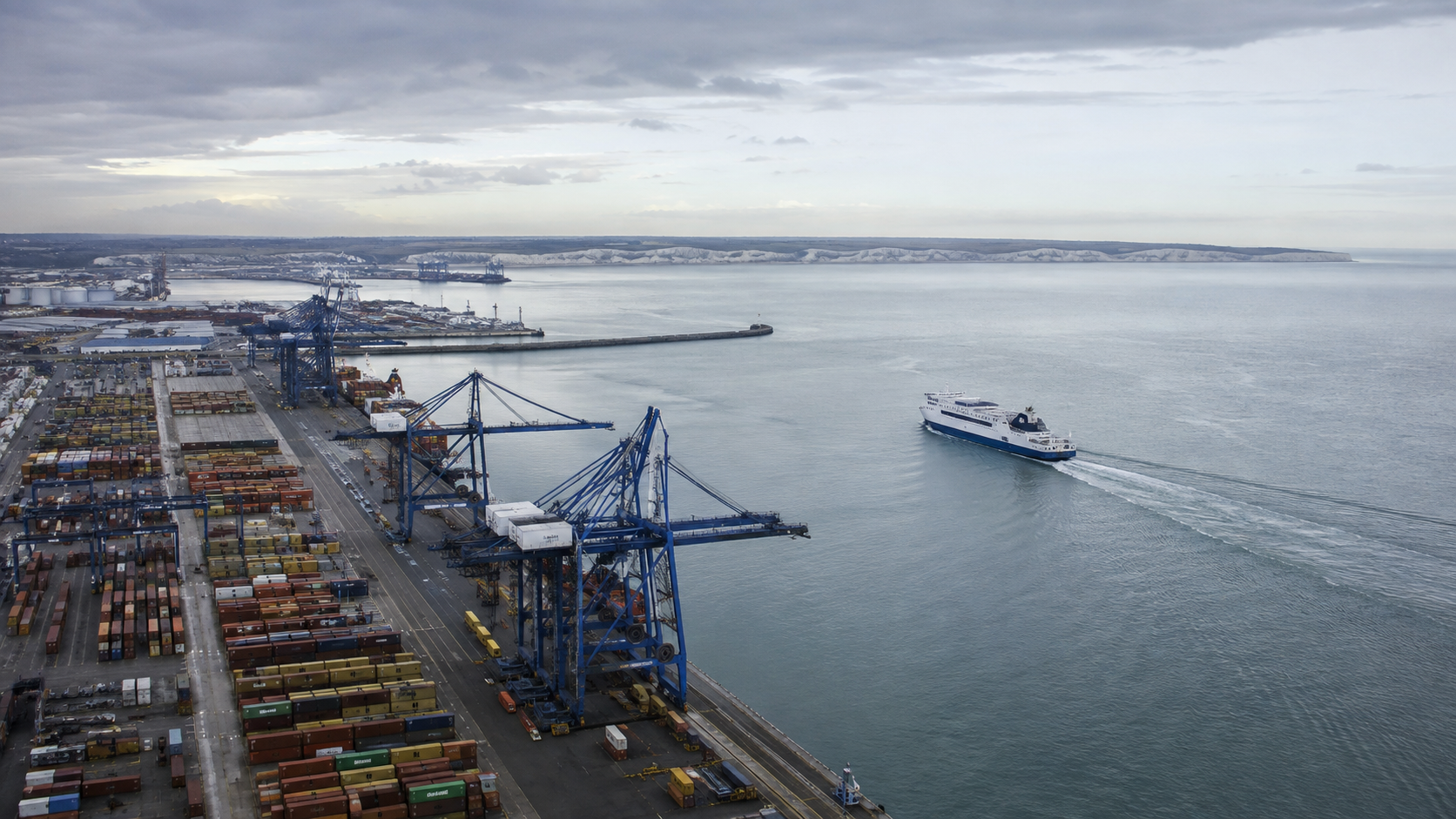 Vue aérienne d'un port de conteneurs avec grues et d'un ferry traversant la Manche, mer grise et calme.