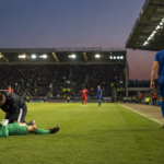 Un gardien de but allongé sur la pelouse reçoit des soins pendant qu'un coéquipier rejoint la ligne de touche pour sortir du terrain, stade éclairé au crépuscule.