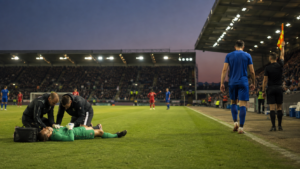 Un gardien de but allongé sur la pelouse reçoit des soins pendant qu'un coéquipier rejoint la ligne de touche pour sortir du terrain, stade éclairé au crépuscule.