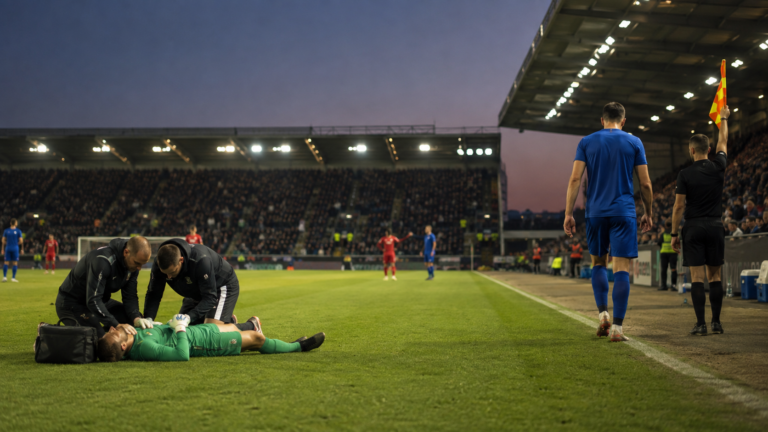 Un gardien de but allongé sur la pelouse reçoit des soins pendant qu'un coéquipier rejoint la ligne de touche pour sortir du terrain, stade éclairé au crépuscule.