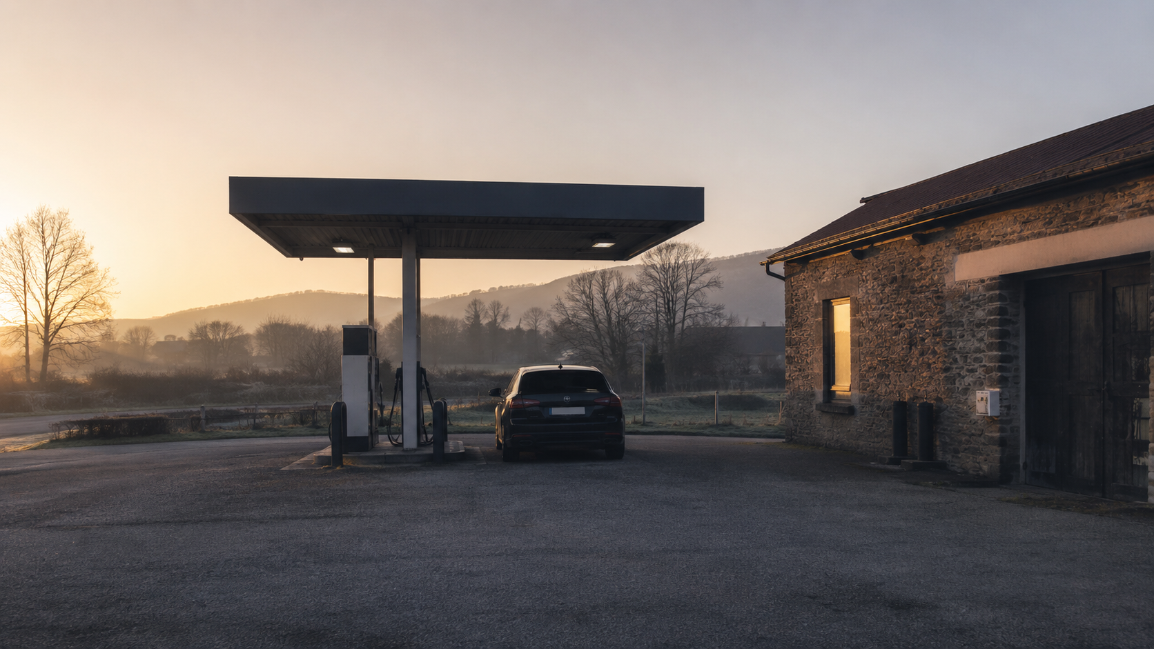 Petite station-service rurale à l'aube, deux pompes sans marque visible, cour vide et voiture stationnée vue de dos.