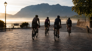 Cyclistes équipés de sacoches s’éloignant d’une place au bord d’un lac alpin au lever du jour, sans véhicule d’assistance