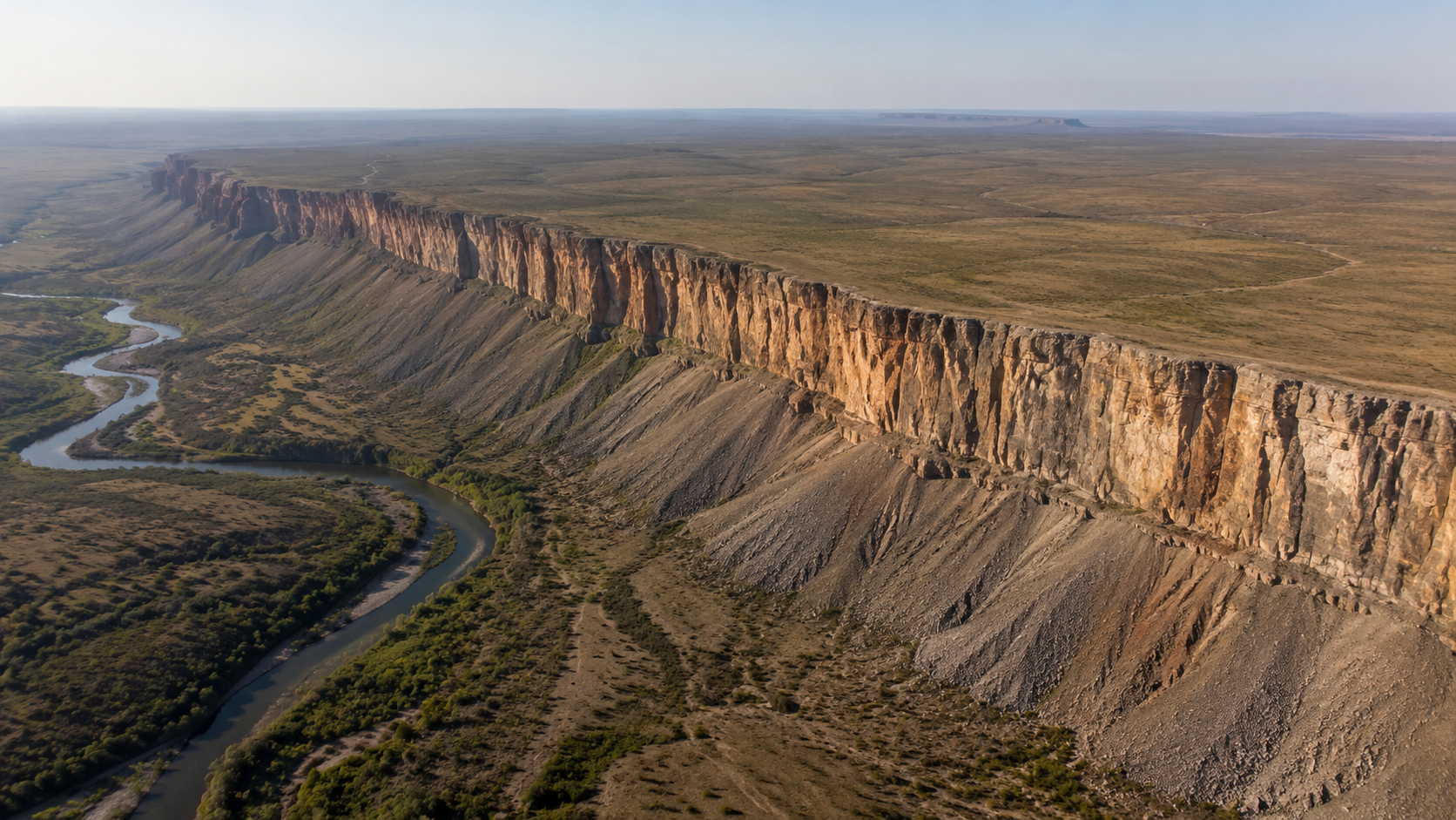 Vue aérienne d’un long escarpement continu haut de plusieurs centaines de mètres traversant un paysage aride, avec des rivières entaillant le plateau et des cônes de débris à son pied.