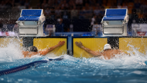Deux nageurs touchent la plaque d’arrivée presque en même temps dans un bassin olympique couvert, gerbes d’eau et tribunes floues en arrière-plan.