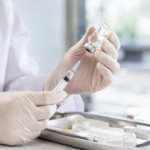 Gros plan sur les mains gantées d'une soignante préparant une seringue de vaccin sur un plateau, dans un cabinet médical lumineux, sans visage reconnaissable.