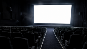 Salle de projection vide, rangées de fauteuils sombres tournées vers un écran blanc allumé.