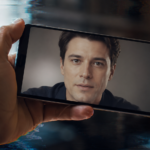 Une main tient un smartphone affichant une vidéo en pause d'un visage générique un peu trop lisse, avec de légères déformations numériques autour des doigts.