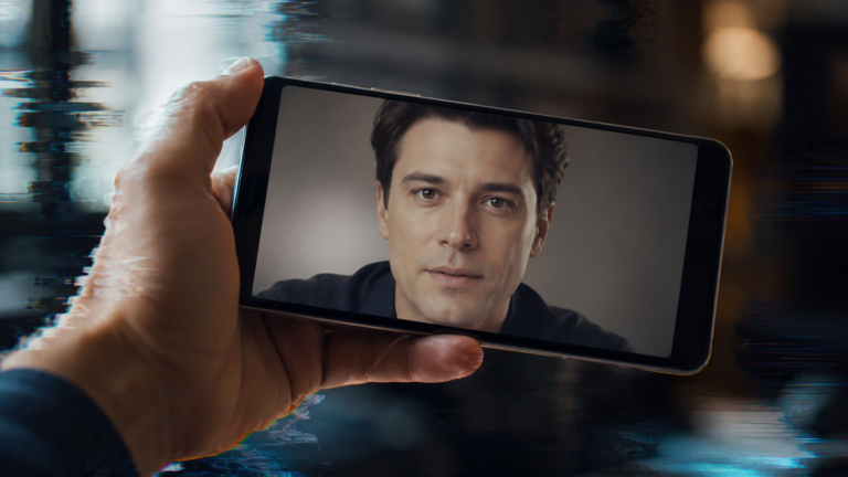 Une main tient un smartphone affichant une vidéo en pause d'un visage générique un peu trop lisse, avec de légères déformations numériques autour des doigts.