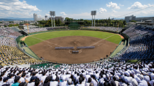 Vue depuis les hauteurs des tribunes d'un stade de baseball japonais comble, terrain en terre battue et herbe sous le soleil d'août