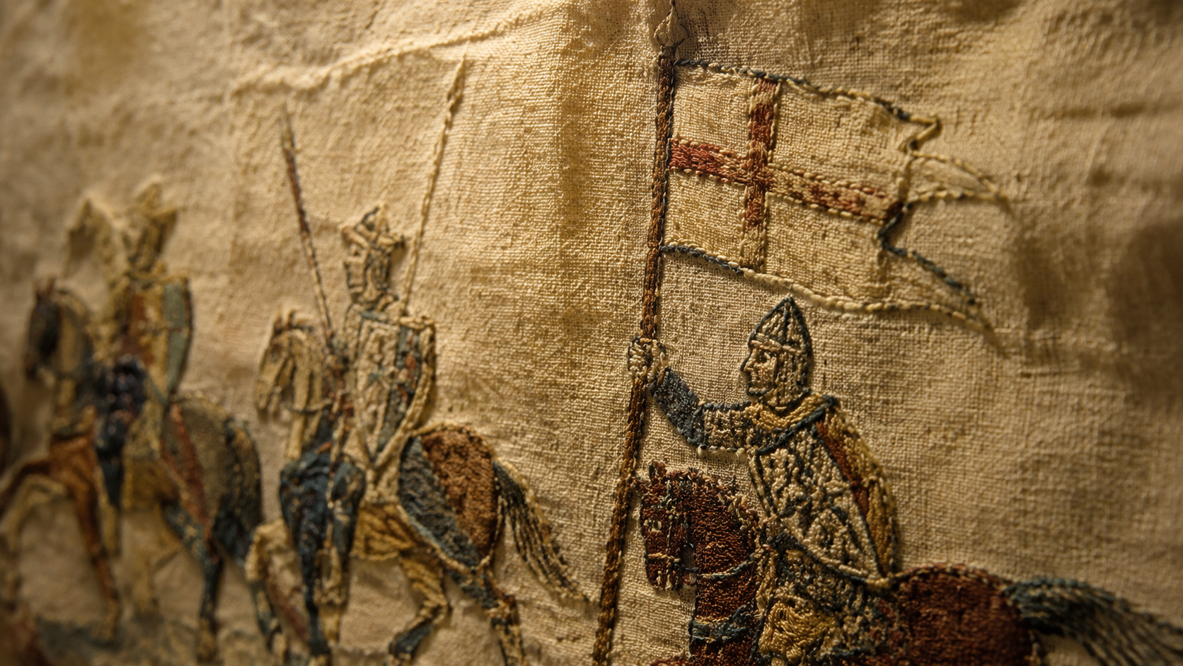 Détail d’une broderie médiévale en lin montrant des cavaliers en armes et une bannière à croix portée sur une hampe.