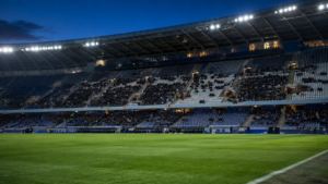 Tribunes partiellement remplies d'un stade de football éclairé à la tombée du jour, rangées vides visibles entre les groupes de supporters, avant le coup d'envoi.