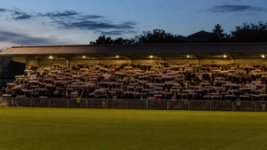 Tribune compacte et bondée d’un petit stade de football de province, écharpes levées sous les projecteurs à la tombée du jour.