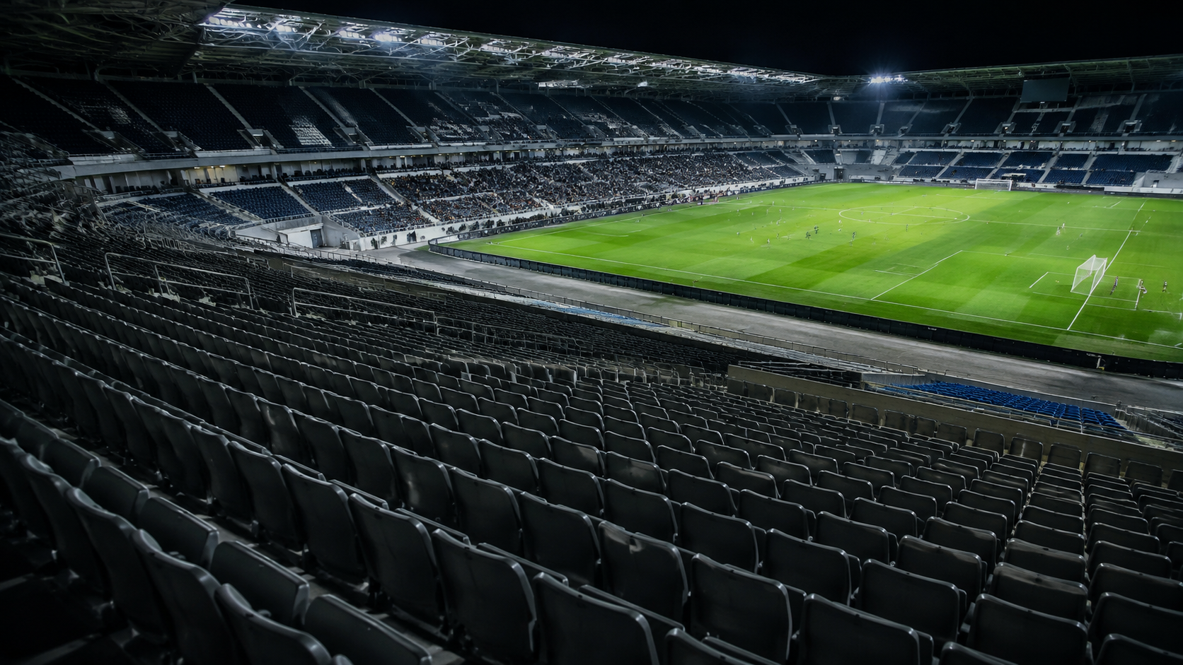 Longues rangées de sièges vides au premier plan d’un grand stade de football pendant un match en nocturne.