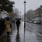 Rue londonienne sous un ciel gris et bas, trottoir mouillé qui reflète la lumière, quelques passants de dos tenant des parapluies dans une bruine fine.