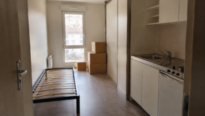 Studio étudiant vide avec un lit sans matelas, une petite kitchenette et des cartons de déménagement empilés près de la fenêtre.