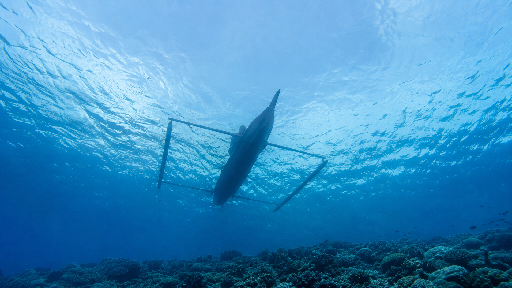 Silhouette d’une petite pirogue à balancier indonésienne vue depuis le fond, à travers l’eau bleue au-dessus d’un récif