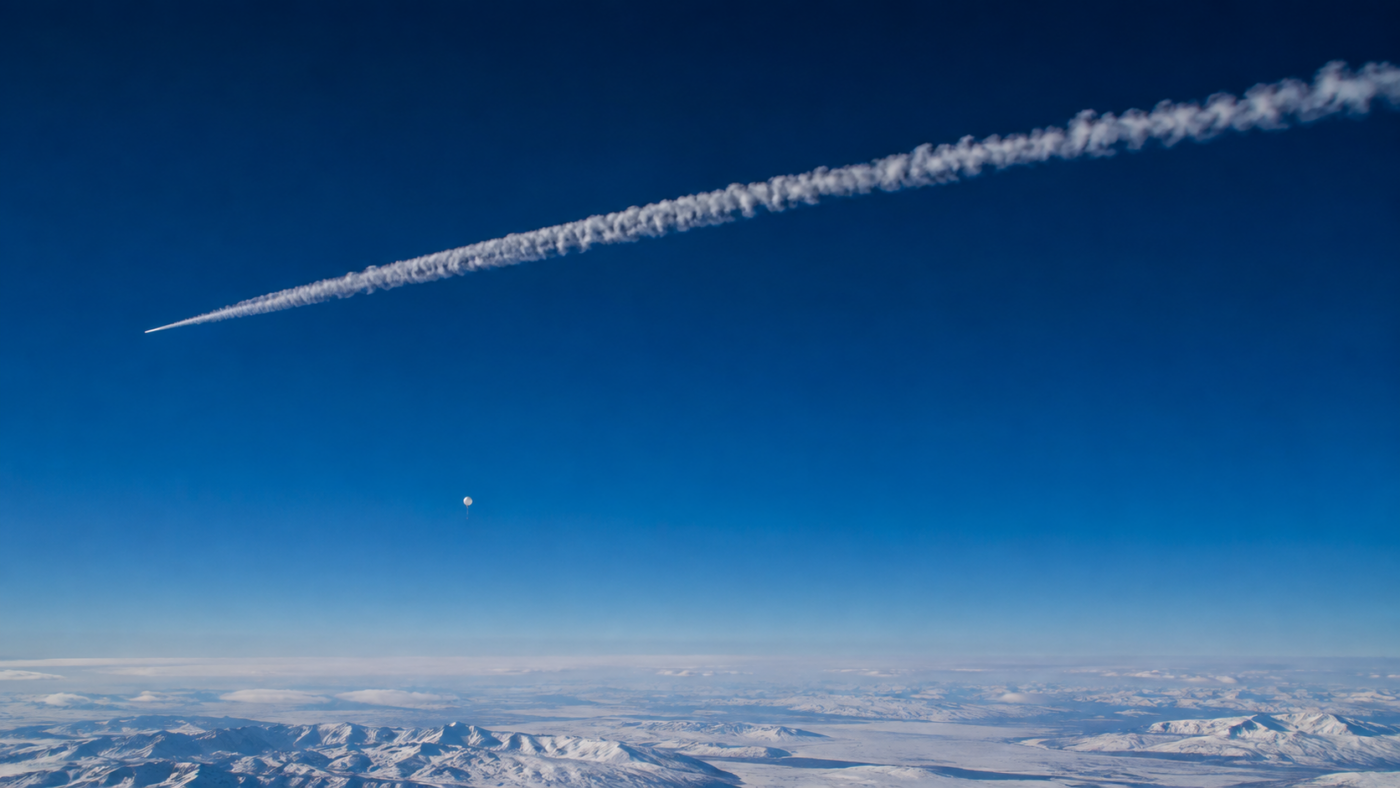 Traînée de condensation blanche dans un ciel d’altitude au-dessus d’un horizon enneigé, avec un minuscule ballon clair en contrebas