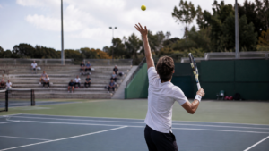 Un jeune joueur de tennis vu de dos, prêt à servir sur un court annexe en dur aux tribunes clairsemées