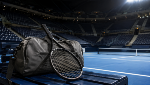 Court en dur bleu éclairé de nuit dans un grand stade, une raquette et un sac posés sur le banc de touche, tribunes vides floues en arrière-plan.