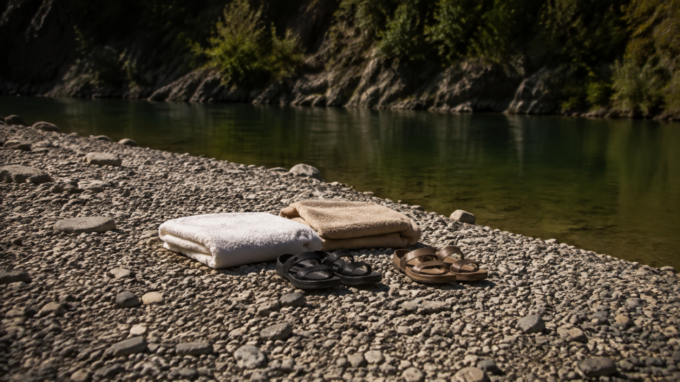 Berge de galets déserte au bord d’une rivière verte, deux serviettes pliées et des sandales posées sur les pierres, sans personne dans le cadre.