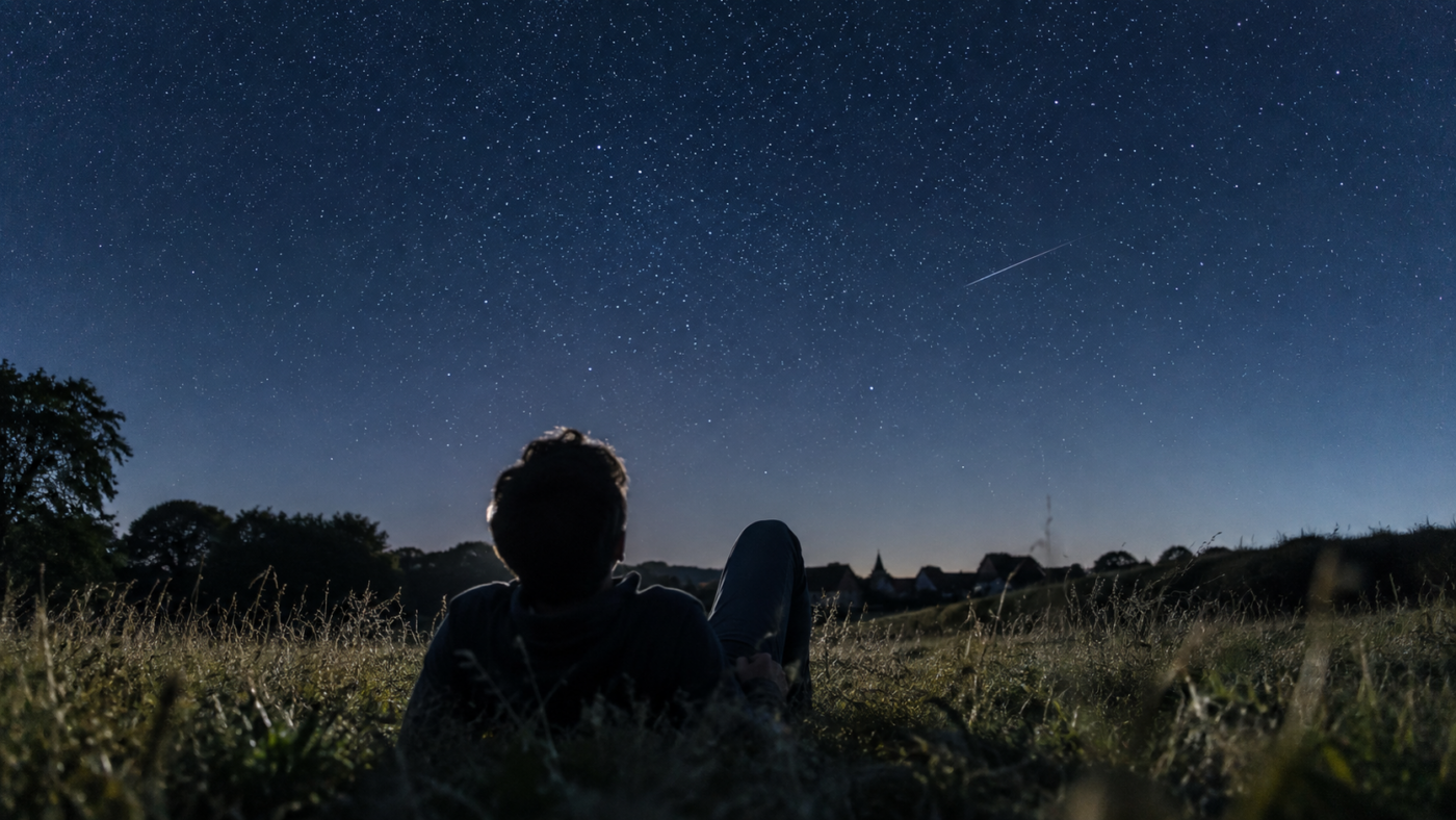 Une personne allongée de dos dans un pré regarde un ciel étoilé dense, sans instrument d’observation.