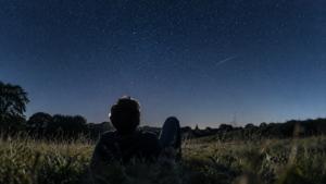 Une personne allongée de dos dans un pré regarde un ciel étoilé dense, sans instrument d’observation.