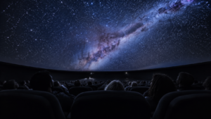 Intérieur d’un planétarium vu du fond de la salle : des silhouettes de dos dans des fauteuils inclinés sous une coupole couverte d’étoiles, aucun visage visible.
