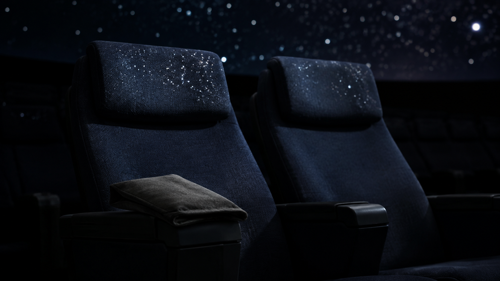 Deux fauteuils inclinés vides dans un planétarium plongé dans le noir, une pochette en tissu fermée posée sur un accoudoir, la lumière des étoiles projetées se reflétant sur les appuie-tête.